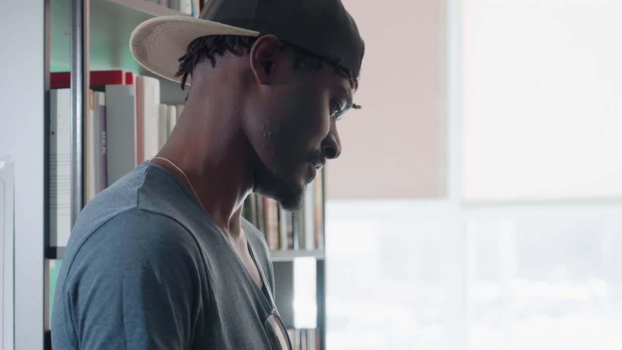 Vista lateral de um homem perto de uma estante em uma biblioteca iluminada, segurando um livro aberto, vestindo camisa casual e boné, lendo atentamente sob a luz natural com um plano de fundo desfocado de livros e luz da janela