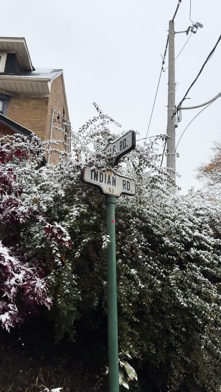 Fixed shot of a snow-covered street sign on Indian Road in Toronto, with winter foliage, a brick house, and a utility pole in the background