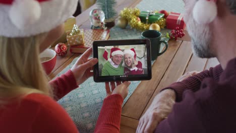 Kaukasisches-Paar-Mit-Weihnachtsmützen-Nutzt-Tablet-Für-Weihnachtsvideoanruf-Mit-Paar-Auf-Dem-Bildschirm