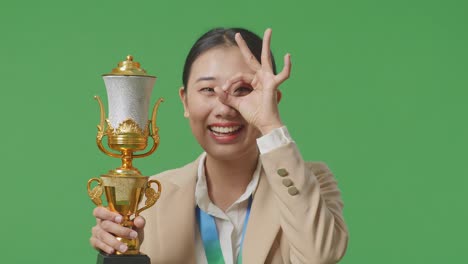 close up of asian business woman in a suit with a gold medal and trophy showing okay hand sign over eye and smiling to camera as the first winner on green screen background in the studio