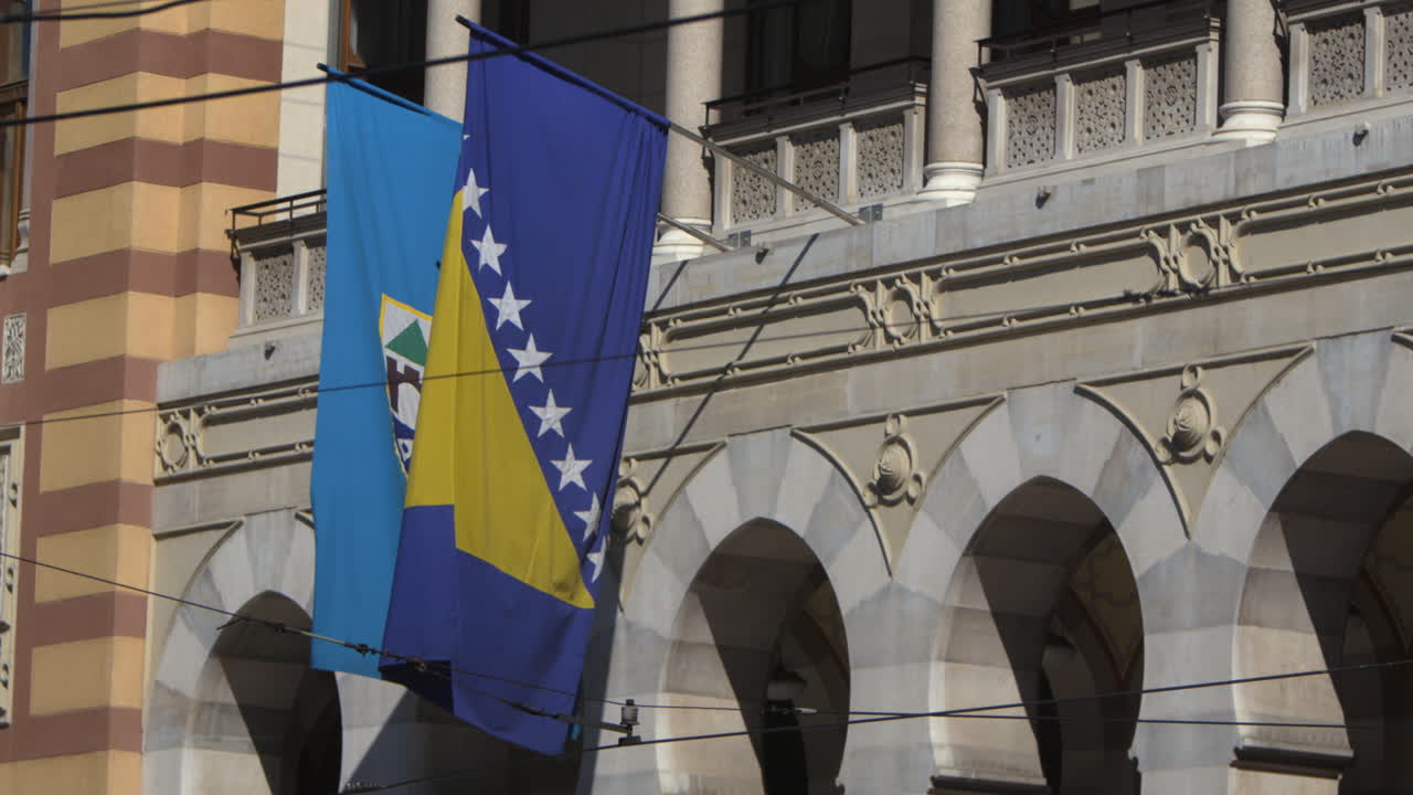 Bosnia and Herzegovina flag blowing in the wind after camera tilt up from bus and pedestrians on street. Seen in Sarajevo capital.