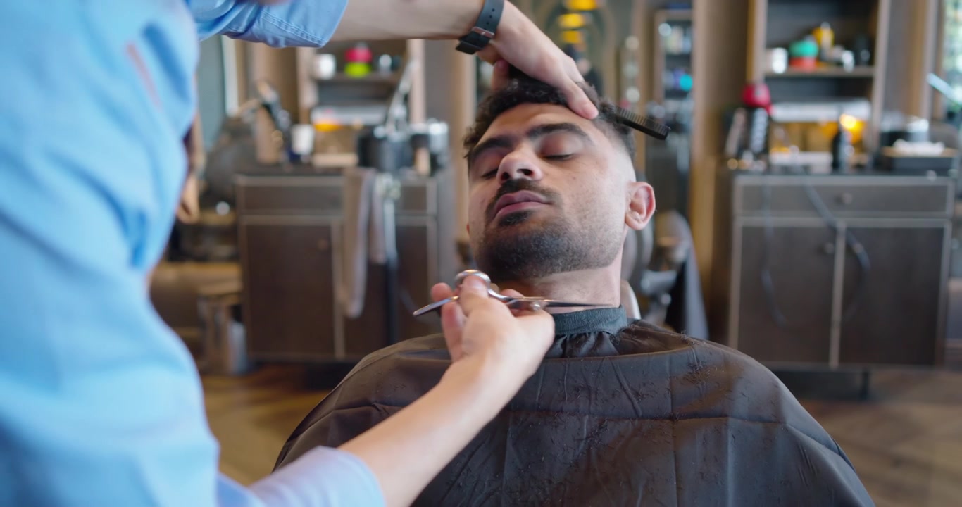 Mandlig barber og kunde læner sig tilbage i barberstolen, barber trimmer skæg med en trimmer i barbersalonen.
