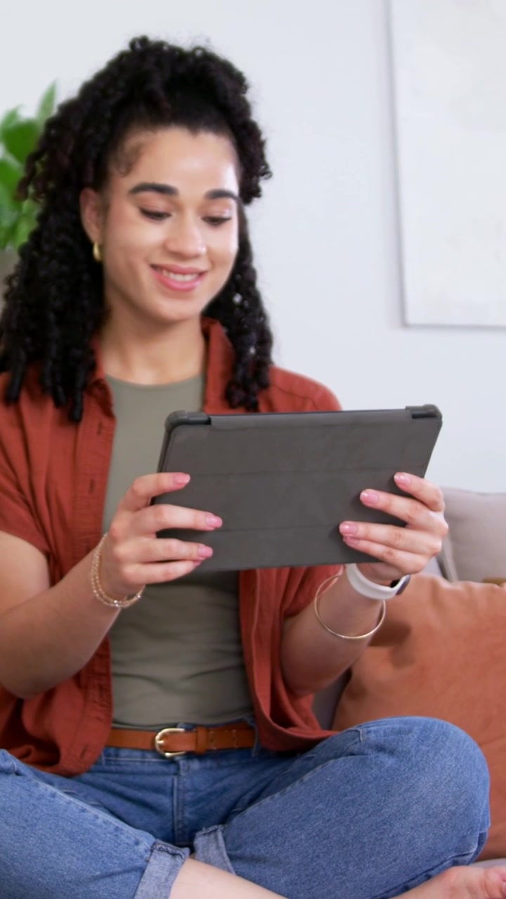 Vertical video: Adjusting, woman in rust top using tablet on sofa as feed appears, camera panning