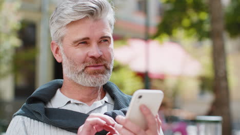 Mature-man-using-smartphone-typing-text-messages-internet-social-media-web-app-looking-up-thinking