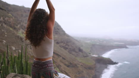 En-Cámara-Lenta,-Una-Mujer-Abraza-La-Postura-Guerrera-Del-Yoga-Junto-Al-Océano,-La-Playa-Y-Las-Montañas-Rocosas,-Capturando-Motivación,-Inspiración-Y-Conexión-Entre-El-Fitness-Y-La-Naturaleza-En-Una-Vida-Saludable.