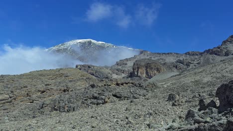 panning shot of rocky mount kilimanjaro hillside with a snow-capped summit rising against a clear blue sky, capturing the rugged volcanic terrain and majestic mountain peak