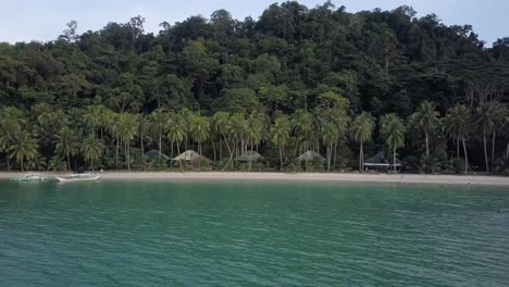 Vista-Aérea-Sobre-Aguas-Turquesas-Hacia-Una-Isla-Tropical-Verde-Con-Palmeras-Y-Cabañas-En-Filipinas---Seguimiento-De-Cámara