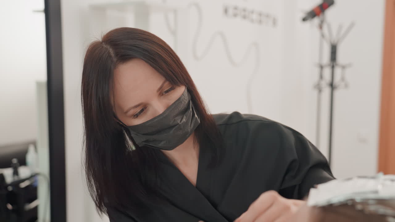 Hvid frisør klipper brunt hår iført maske, omhyggeligt saksearbejde i moderne salon, sort uniform, fokuseret udtryk, spejl og stylingværktøjer synlige, ren belysning, professionel atmosfære
