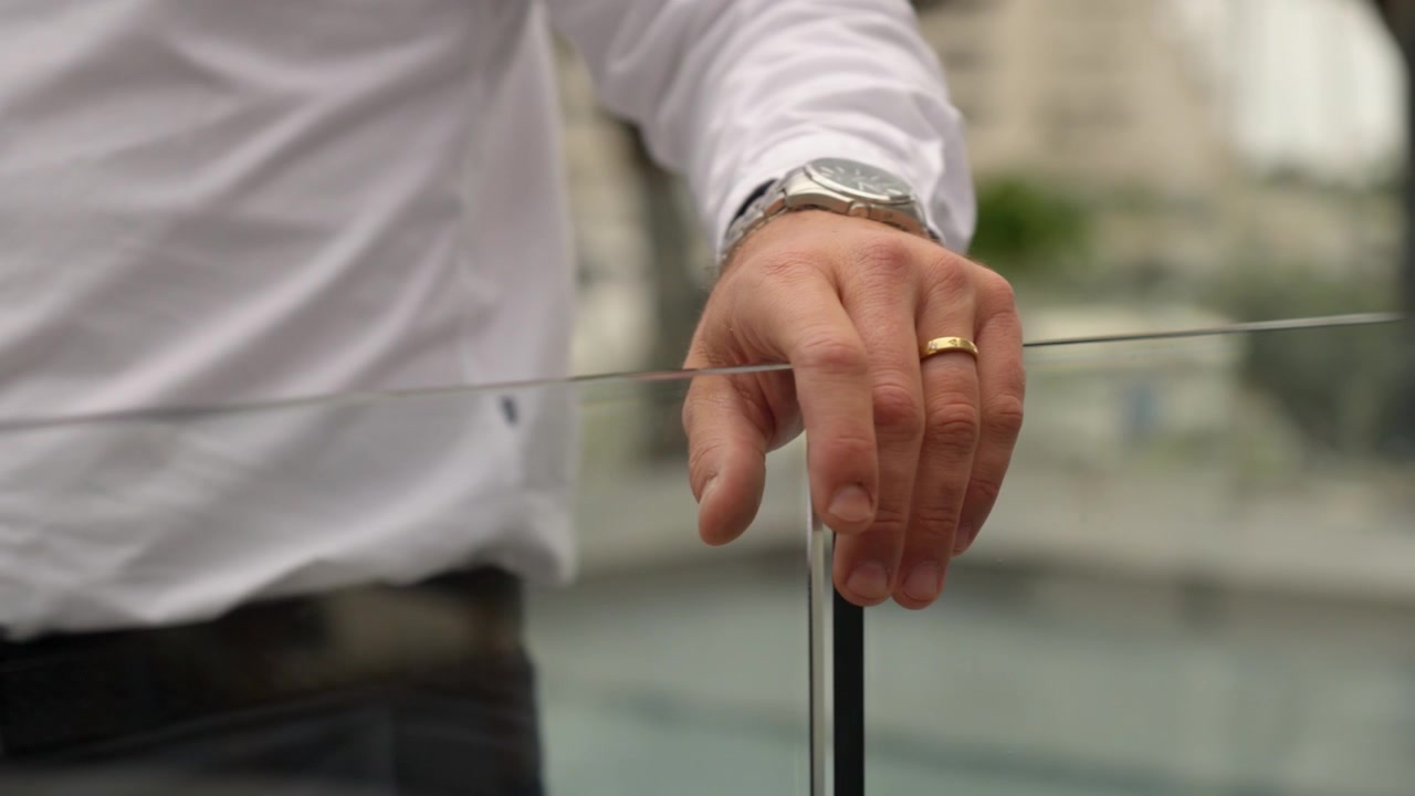 A close-up shows a person in a white shirt handling the top edge of a glass pool gate, with a wristwatch and ring visible against a softly blurred background.