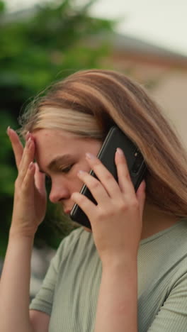 primer plano de una mujer joven que parece agotada durante una llamada telefónica, sosteniendo su mano izquierda en su frente en frustración, el fondo muestra vegetación borrosa y un edificio