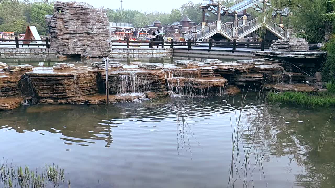 A serene park scene with a man-made waterfall cascading over rocks, and a wooden walkway and building in the background