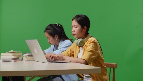 asian woman student having a headache while sitting with her friend and typing on a laptop on the table in the green screen background classroom