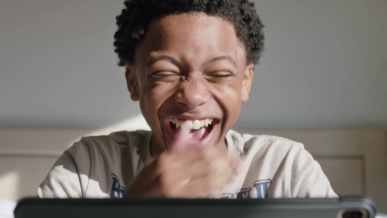 Laughing adolescent male watching funny clip on tablet in bedroom, wearing light T-shirt