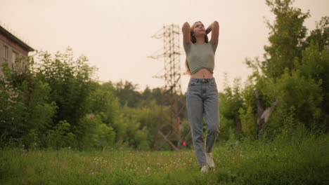 young woman walking on grassy path with hand resting on head, surrounded by lush greenery and distant power lines, a red light and passing car blur in the background