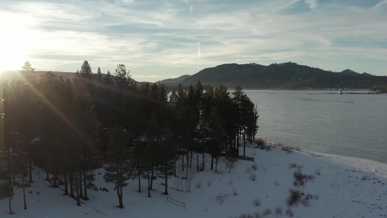 Aerial flyover trees on the shore of a frozen lake at sunset. 4K
