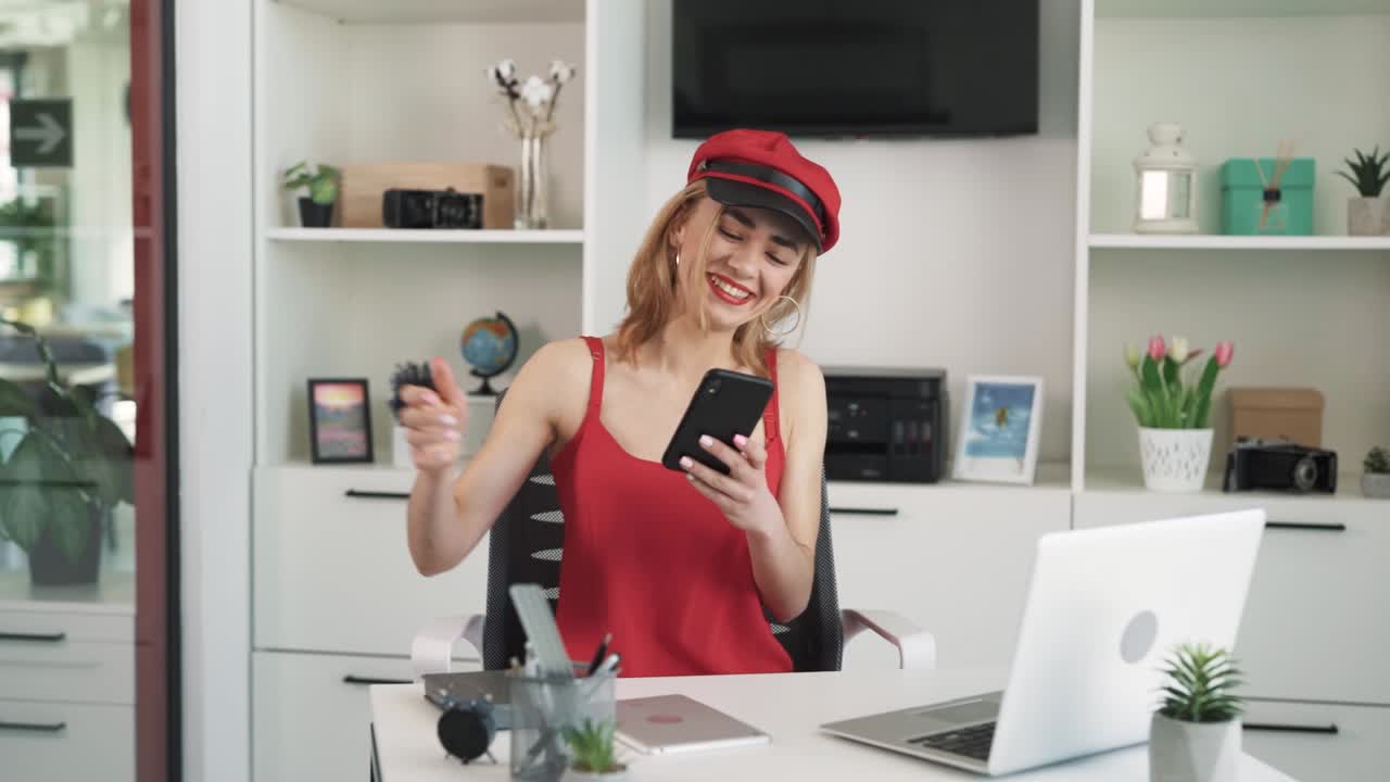 The young woman works in the office with a laptop, she receives a notification on her phone,the woman is surprised and joyful
