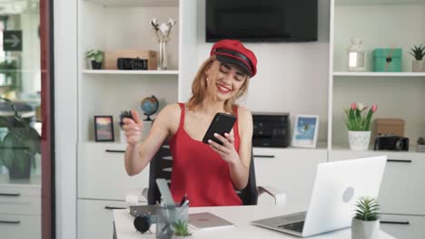 the young woman works in the office with a laptop, she receives a notification on her phone,the woman is surprised and joyful