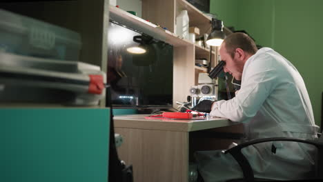 a close view of a scientist in a white lab coat working diligently at a desk in a laboratory, the scientist is using a microscope and a cell phone and is surrounded by various tools