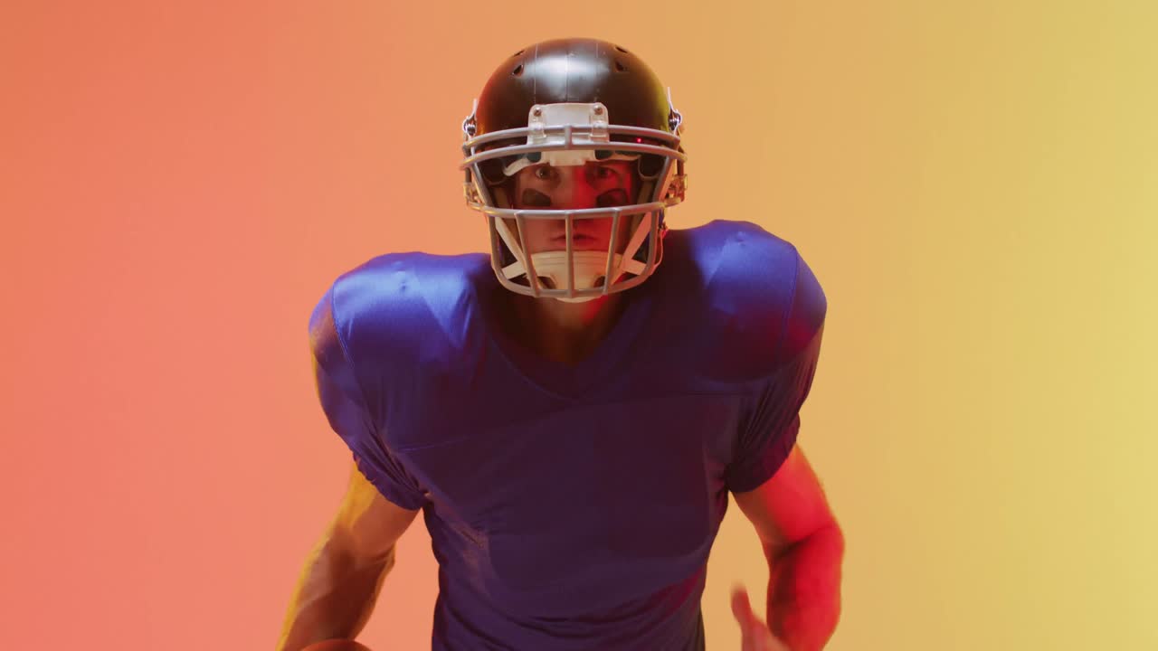 Video ravvicinato di un giocatore di football americano caucasico con casco e pallone su sfondo arancione.