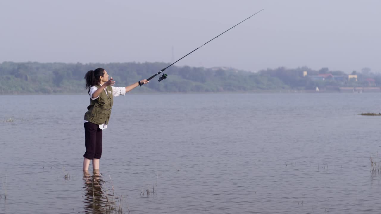 Seorang wanita Asia Selatan berdiri di air danau yang dangkal sambil memegang pancing dan mengamati tepi danau yang tenang, menikmati alam pedesaan, dan ketenangan