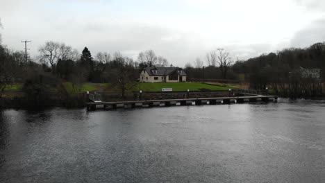 aerial - fly through grass field and a river towards a single english house nearby the lake