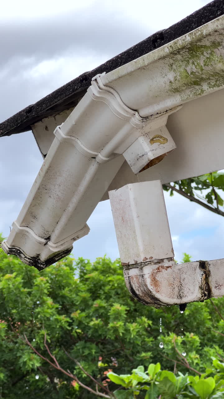 Medium shot af en våd, hvid tagrende med vanddråber, der hænger fra kanten, fastgjort til et mørkegråt tegltag på et beboelseshus. Overskyet stemning efter regn. 4K ProRes 30fps