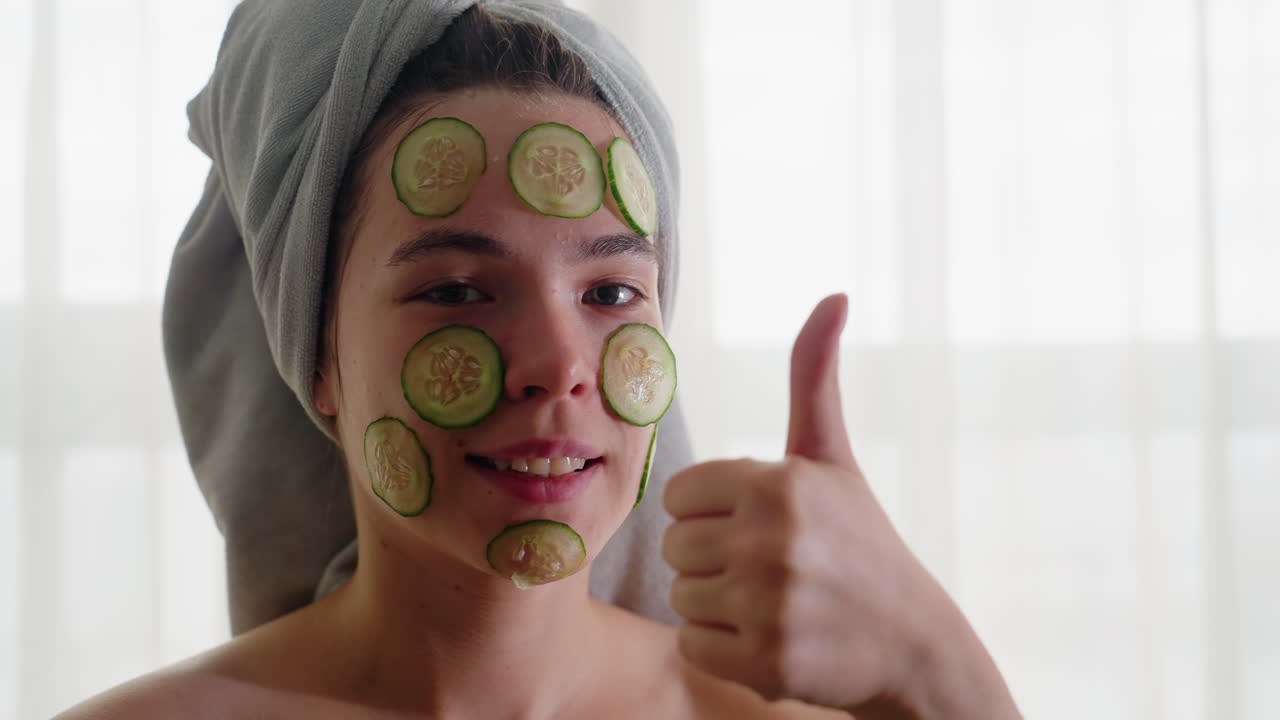 Close-up of young woman smiling with cucumber slices on face, hair wrapped in towel, giving thumbs up while enjoying refreshing skincare and wellness moment during peaceful self-care beauty routine