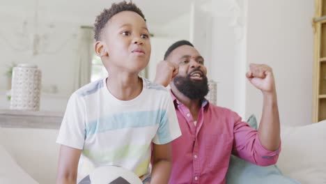 happy african american father and son watching football match and supporting, in slow motion