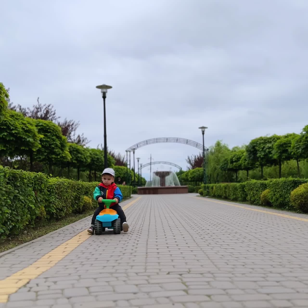 Small kid on the toy car approaches camera by the alley in the park. Cute kid comes close smiling adorably
