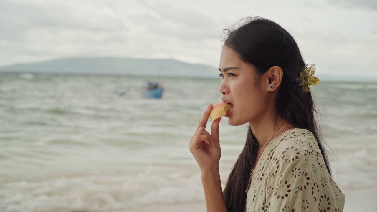 Female enjoys fresh seaside bounty. Woman delicately tastes fresh produce by tranquil shoreline and boat. Lady silently savors juicy fruit while relaxing by seaside with waves and boat nearby
