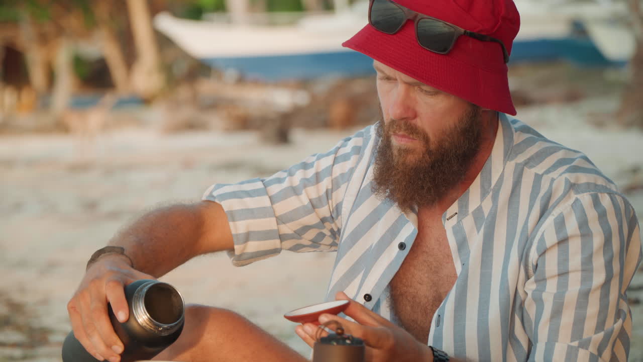Uomo bianco concentrato, da solo in spiaggia, con un cappello da pescatore rosso, versa il caffè dal thermos nella tazza, camicia a righe, profilo barbuto, barche a riva, ora d'oro, rituale del viaggiatore all'aperto, calma solitudine marina.
