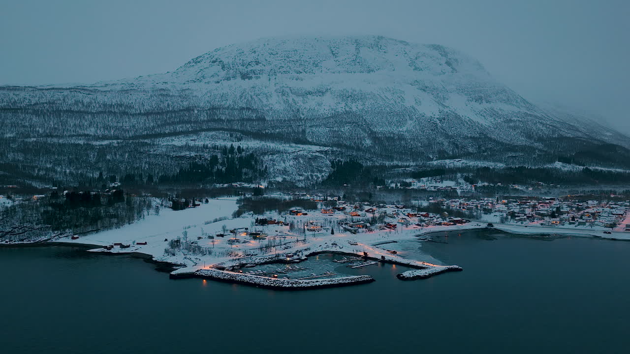 Filmatisk flyfoto av en liten norsk fiskerlandsby idyllisk plassert mellom snødekte fjell og havet under blåtimen. Perfekt for reise- og vinternaturdokumentarer.