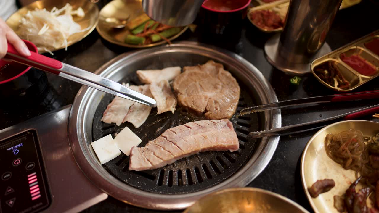 Hand uses tongs to flip pork belly slices on tabletop Korean barbecue grill, warm lighting