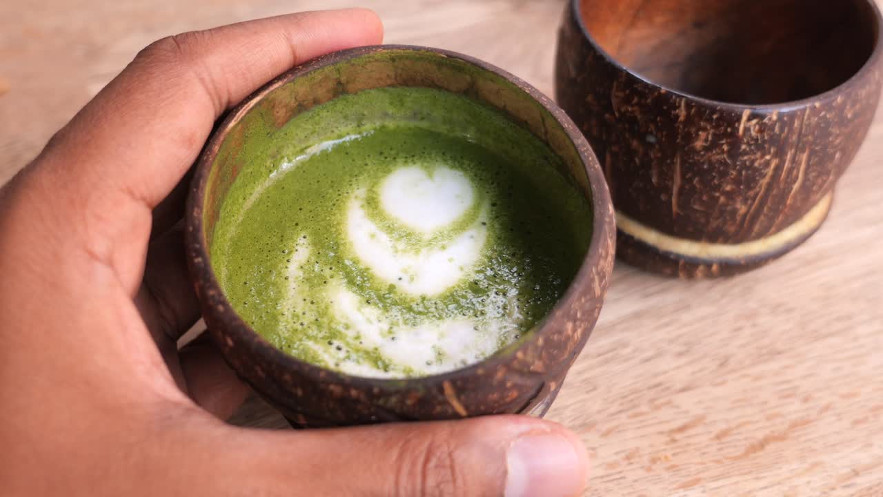 una mano sosteniendo una taza de cáscara de coco llena de arte matcha latte