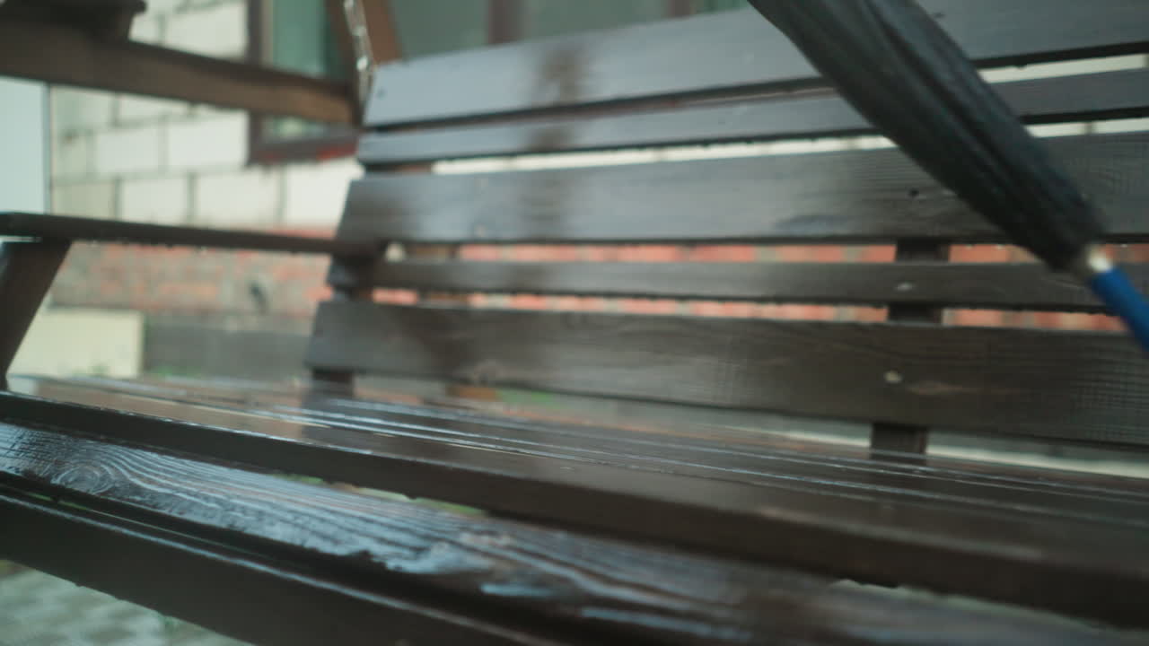 초록색 레인코트를 입은 개인이 나무로 된 스 벤치에 놓인 은 우산을 잡으려고 손을 는 클로즈업, 배경에는 흐릿한 건물이 있습니다.