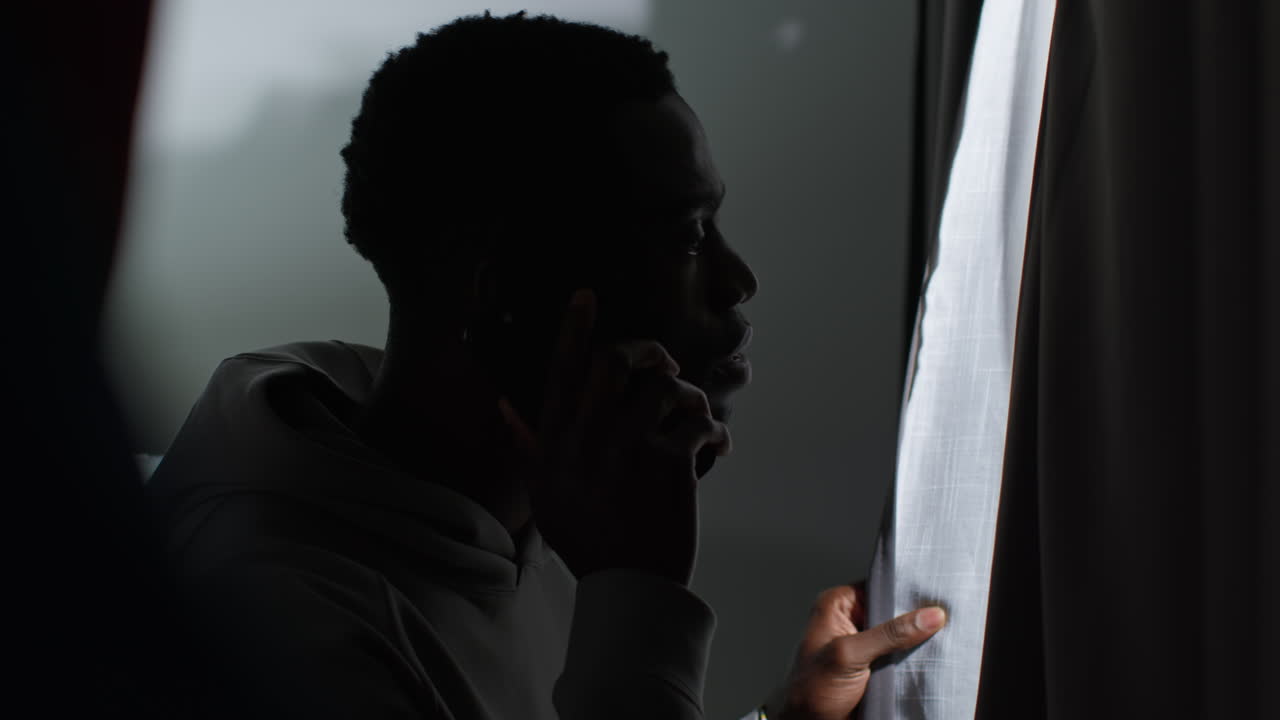 Young Man With Mental Health Issues Suffering With Paranoia Or Agoraphobia At Home Talking On Mobile Phone Looking Out Of Window 1