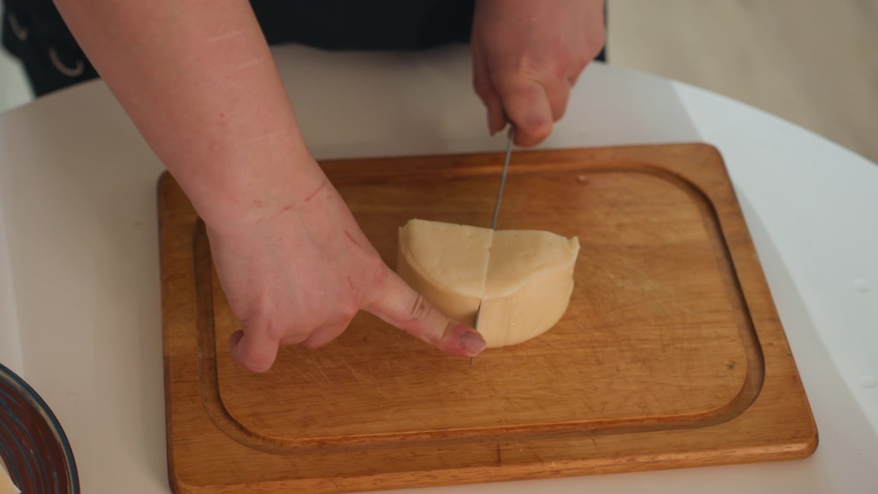 Close-up de mãos cortando queijo redondo em uma tábua de corte de madeira na cozinha de casa, com uma mão segurando o queijo enquanto a outra guia uma faca serrilhada através de fatias lisas e uniformes para cozinhar, servir e.
