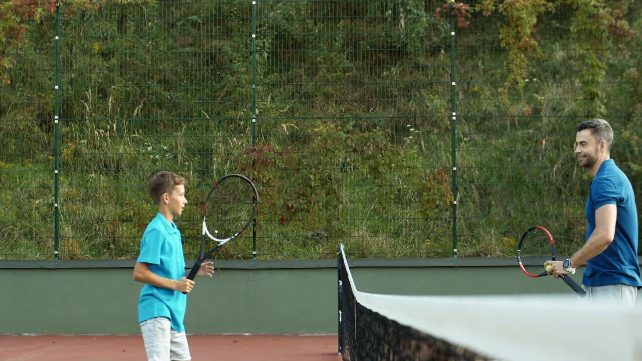 아버지와 10대 아들이 스포츠 코트에서 야외 테니스 경기 전후에 웃고 환호하는 모습