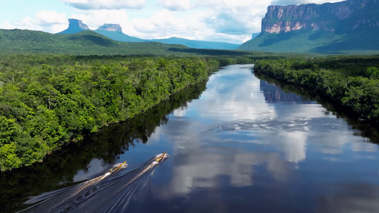 Two boats navigate a vast river through a lush rainforest with majestic tepui mountains in the background