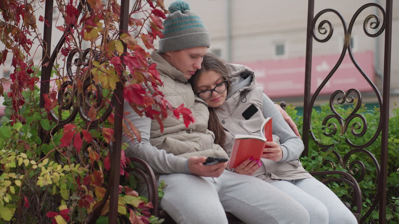 Husband and wife seated outdoor park bench reading book together as wife rests on husband shoulder, man holds phone while gently placing hand on book, autumn leaves sway with the wind