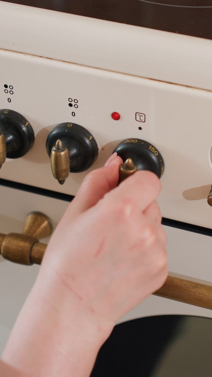 Un primo piano mostra una mano adulta che afferra una manopola di controllo nera su un pannello di un fornello color crema, girandola accanto a una barra di ottone, una spia rossa e un indicatore di temperatura rotondo, catturando un preciso elettrodomestico da cucina