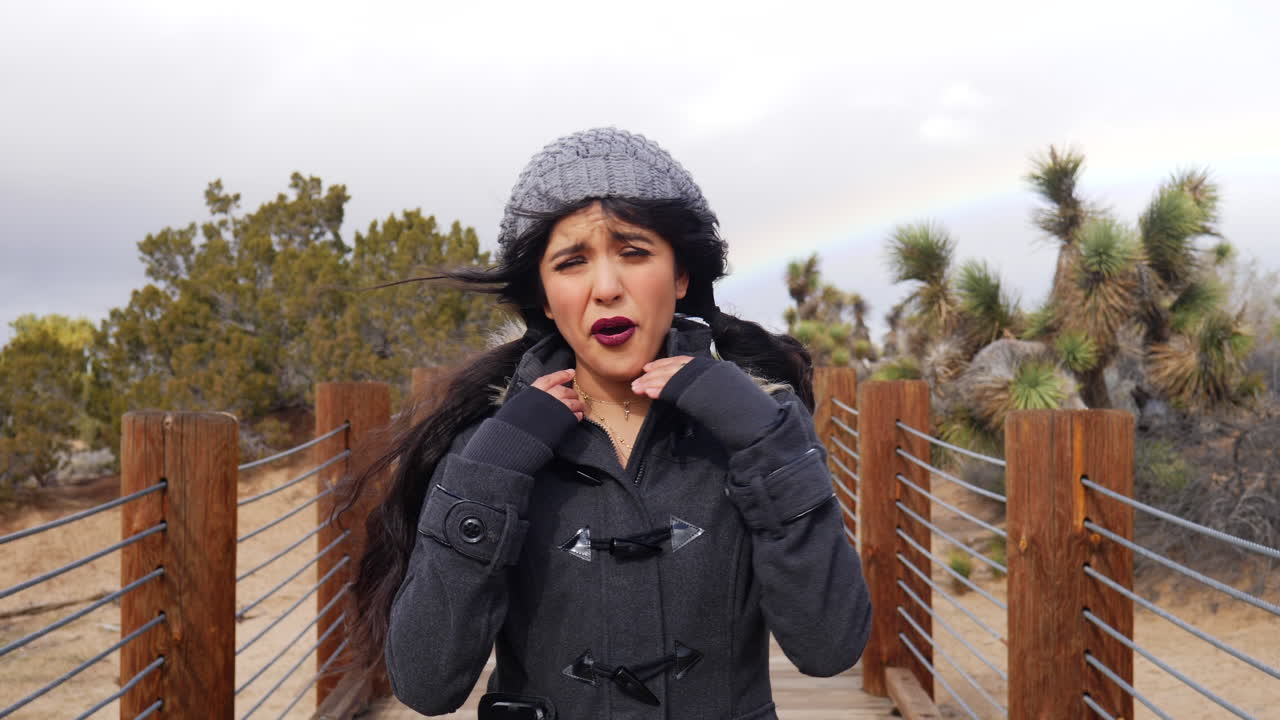 Attractive young woman in a freezing cold and windy storm with a colorful rainbow in the cloudy sky SLOW MOTION