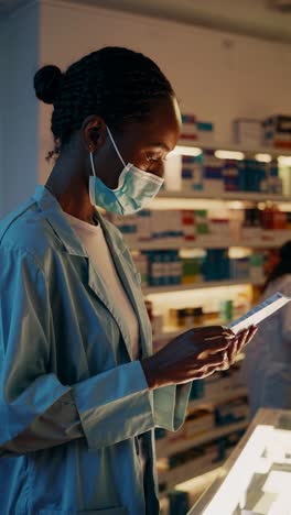 young pharmacist wearing face mask scanning medicine box using smartphone in drugstore, shelves full of pharmaceutical products in background