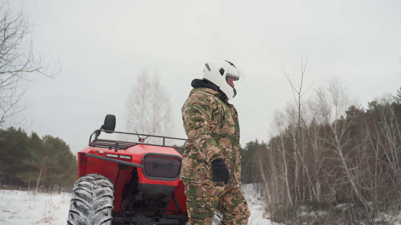 Uomo bianco si allontana dall'ATV inoltrandosi in una foresta di betulle. Un ranger con il casco ispeziona il paesaggio innevato, gli stivali scricchiolano sulla neve e i passi si affievoliscono. Scruta l'orizzonte in cerca di un percorso, una camminata solitaria e contemplativa sotto il grigio inverno.