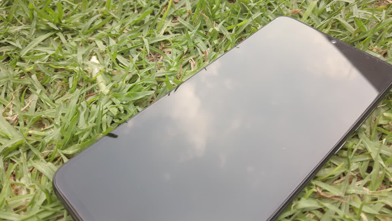 Modern black smartphone lying on green grass reflecting the sky, with slow pan showing reflection and nature contrast.