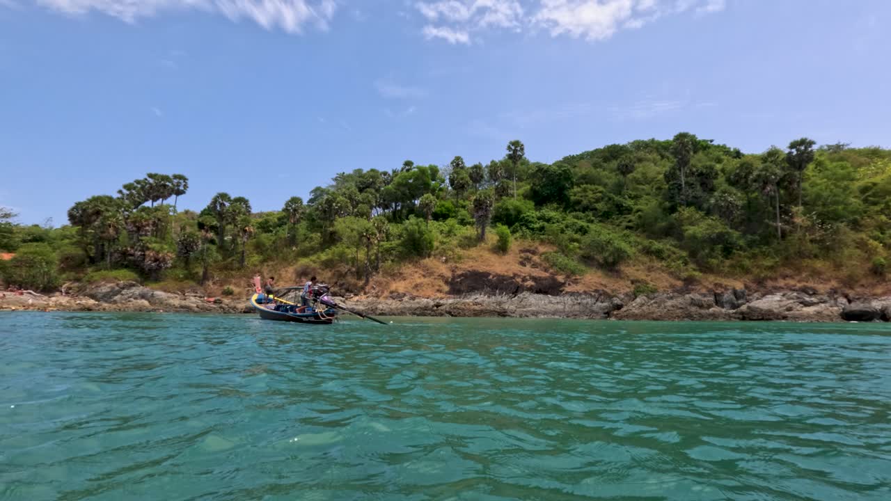 A longtail boat navigates vibrant waters near lush, rocky shores under clear skies in Phuket, Thailand