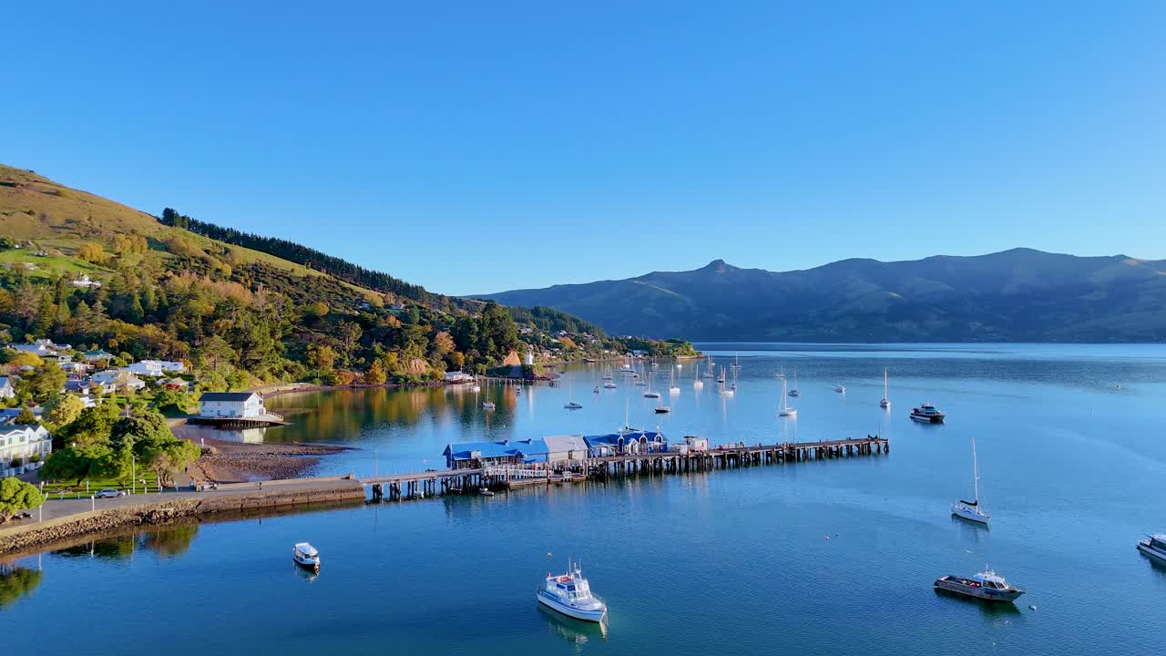 Aerial footage captures Akaroa Harbour's serene waters, boats, and surrounding hills under clear blue skies