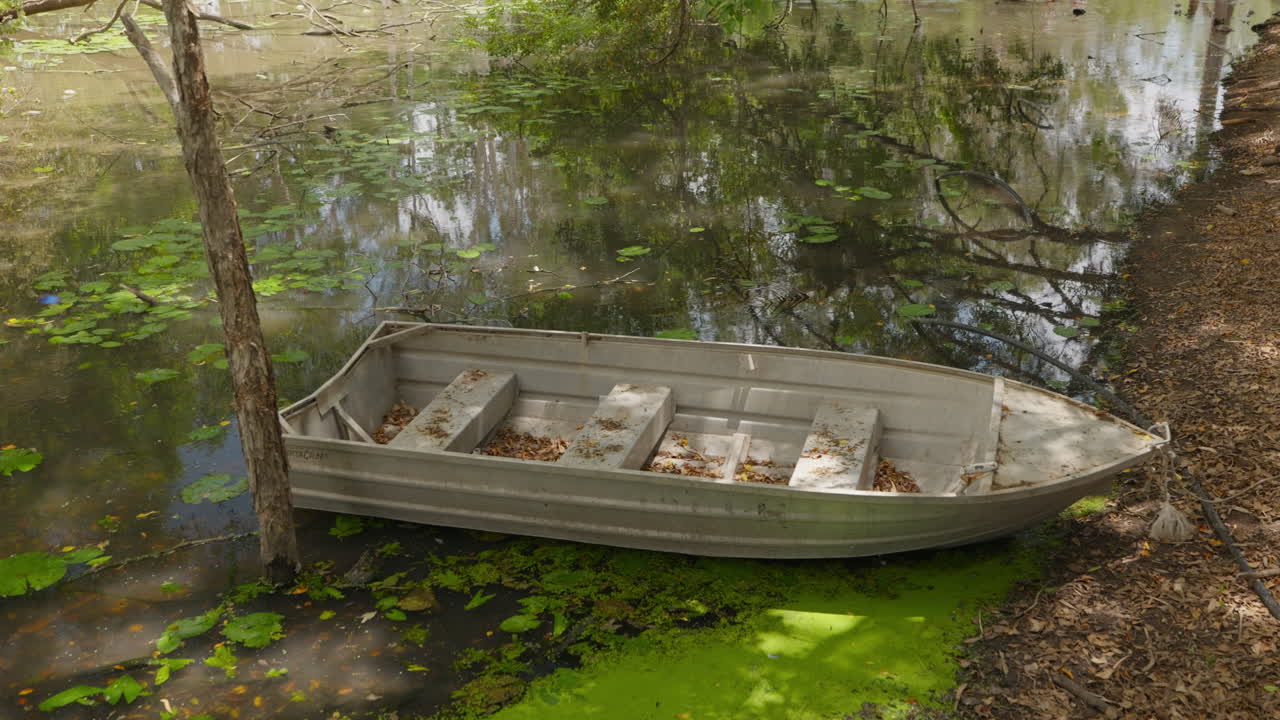 Una barca de aluminio llena de hojas secas que cayeron de los árboles, está atada a la orilla y flotando sobre el agua verde con algas de un lago apartado, con el cielo y los árboles reflejándose en las aguas