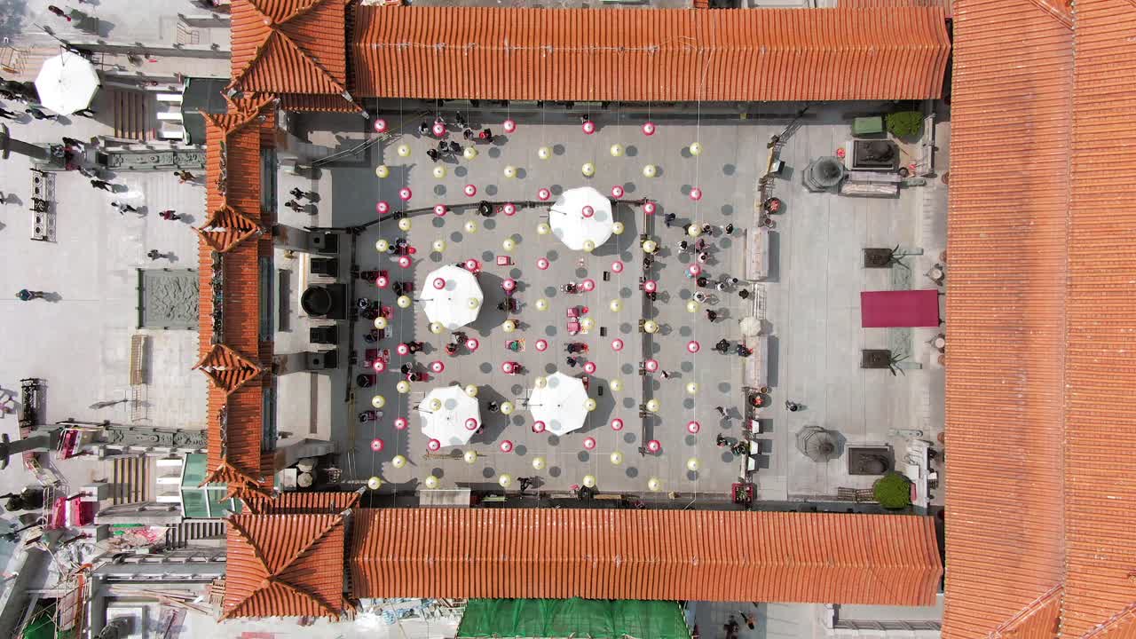 Wong Tai Sin temple court with people lighting incense sticks and kneel before the main altar under Chinese lanterns, Top down one minute aerial view.