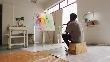 Vista-Trasera-De-Un-Reflexivo-Artista-Afroamericano-Sentado-Mirando-Su-Pintura-En-El-Estudio-De-Arte.